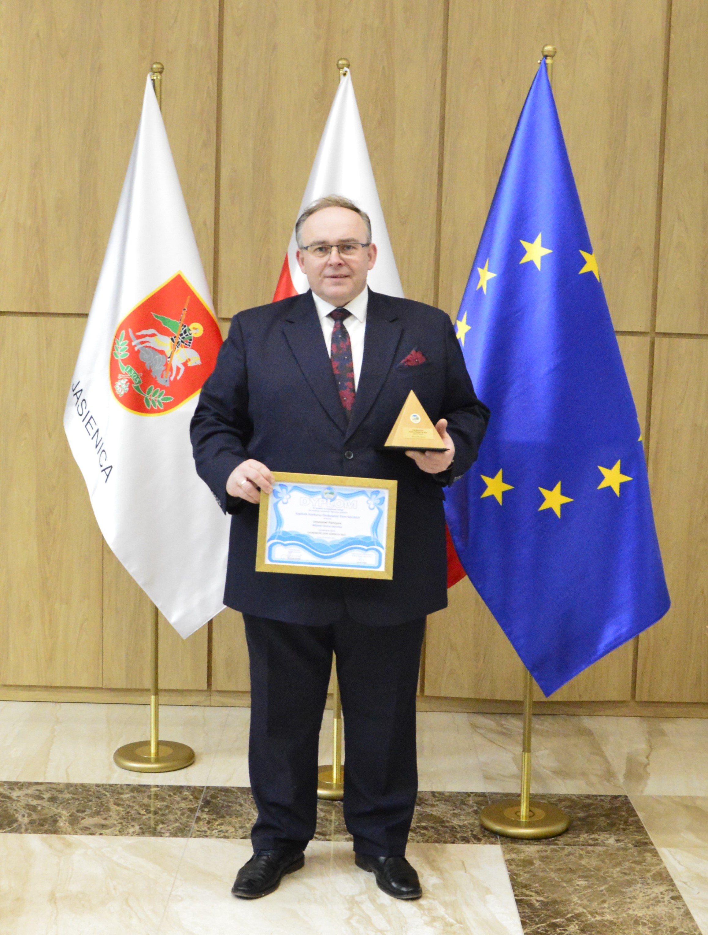 Wójt Gminy Jasienica trzymający dyplom oraz satuetkę Osobowości Ziem Górskich na tle trzech flag - Gminy Jasienicy, Flagi Polski oraz Unii Europejskiej 