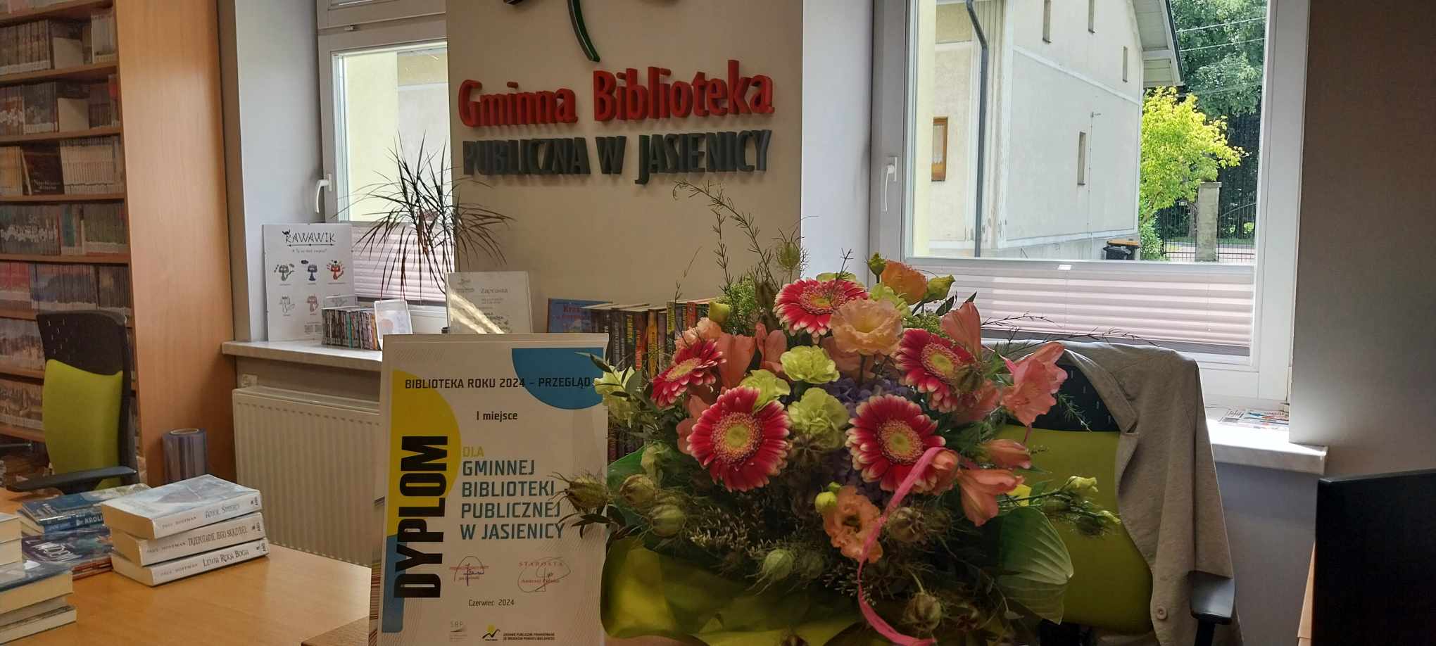Zdjęcie przedstawia w centralnym punkcie dyplom, kwiaty a w tle napis "Gminna Biblioteka Publiczna w Jasienicy", po lewej stronie znajduje się regał z książkami oraz stos książek położony na biurku. 