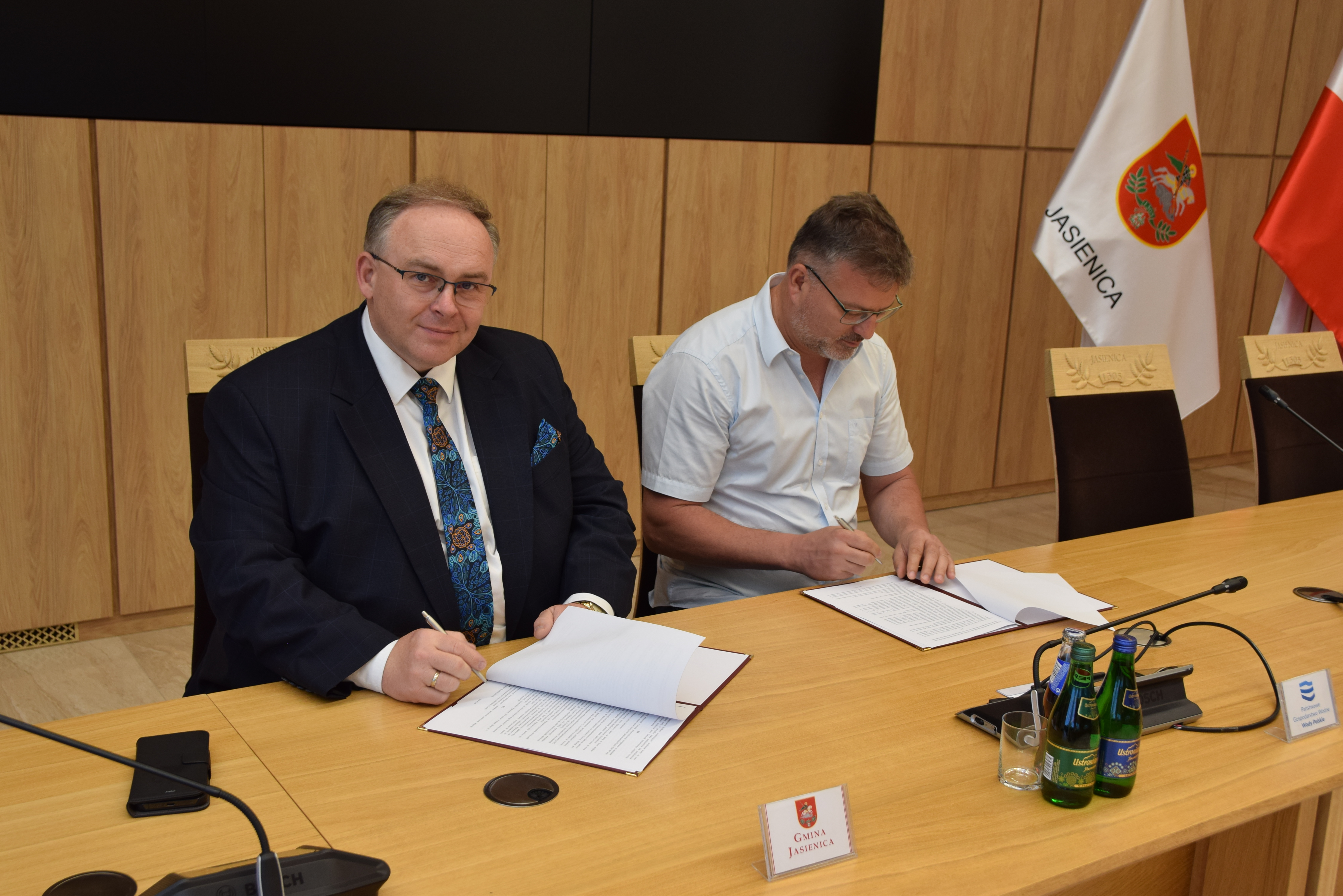 Dwóch mężczyzn siedzi przy stole konferencyjnym w sali obrad. Po lewej Janusz Pierzyna, Wójt Gminy Jasienica, podpisuje dokument i spogląda w stronę obiektywu z lekkim uśmiechem. Po prawej Wojciech Solik podpisuje dokument. W tle widoczne są flagi: gminy Jasienica i Polski. Na stole znajdują się dokumenty, mikrofony, woda oraz tabliczki z nazwami instytucji.