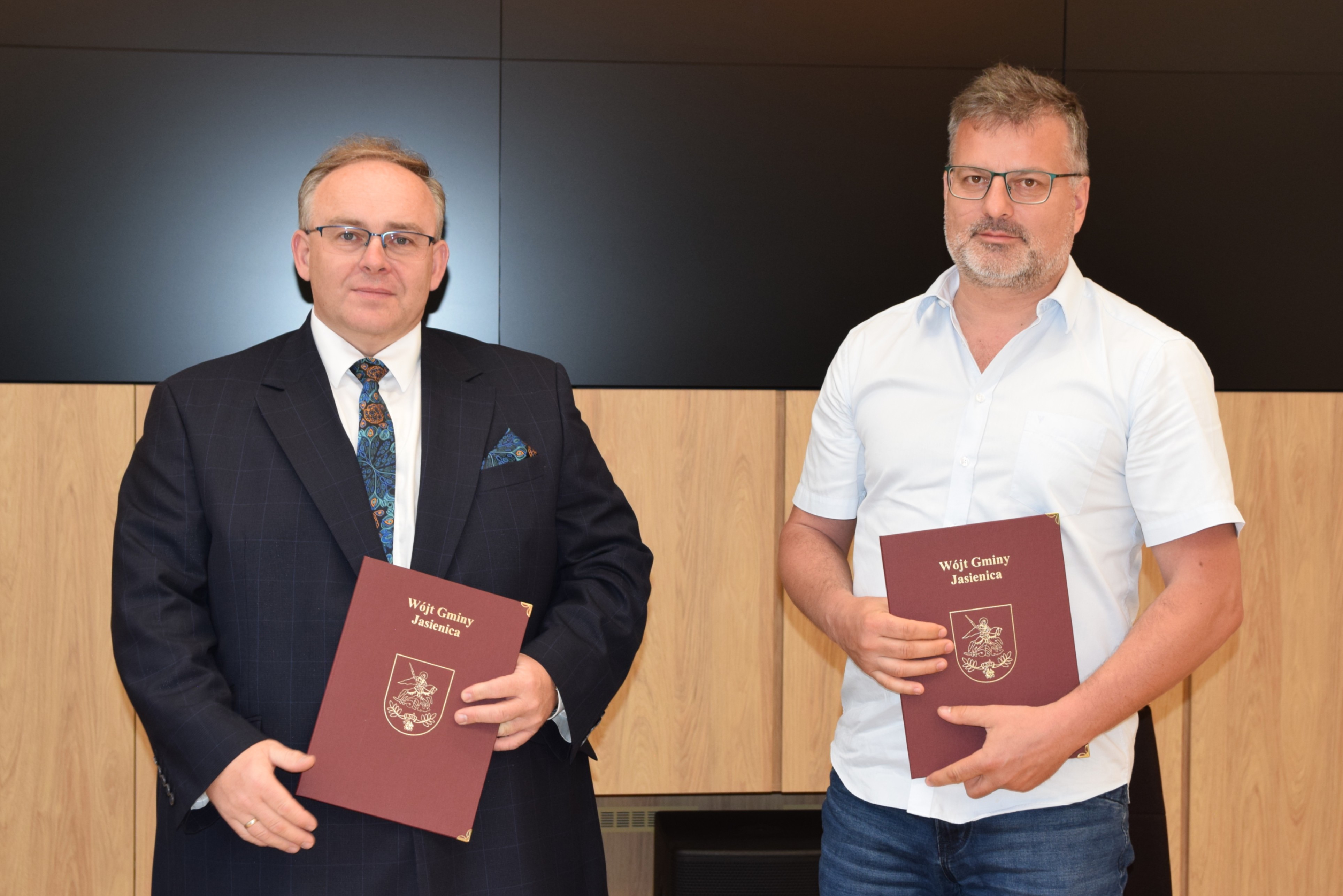Dwaj mężczyźni stoją obok siebie na tle drewniano-czarnej ściany. Po lewej stronie Janusz Pierzyna, Wójt Gminy Jasienica, trzyma bordową teczkę z godłem gminy i napisem „Wójt Gminy Jasienica”. Po prawej Wojciech Solik z „Wód Polskich” trzyma identyczną teczkę. Obaj patrzą w stronę aparatu i pozują do wspólnego zdjęcia po podpisaniu porozumienia.