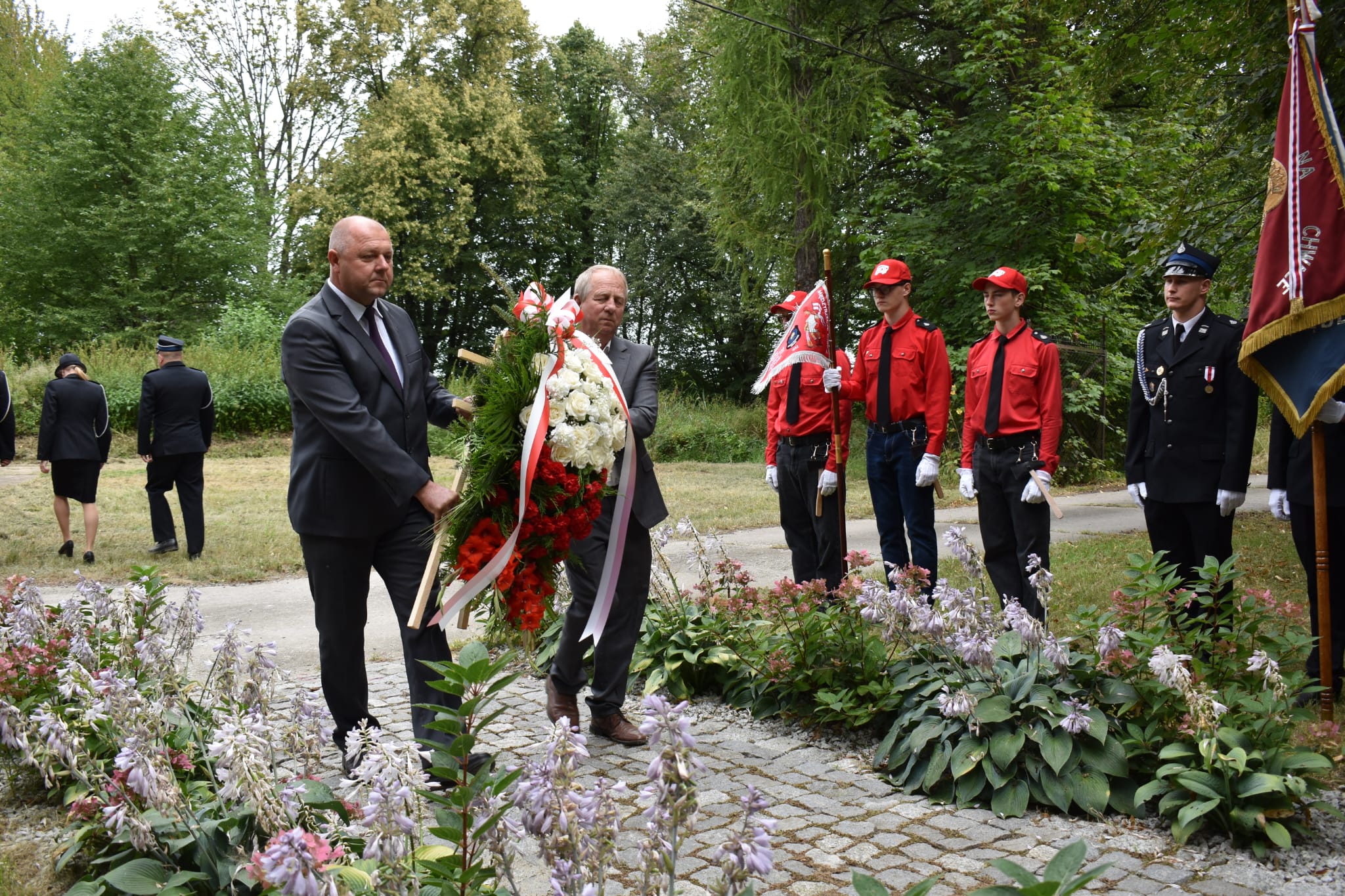 Na zdjęciu widać dwóch mężczyzn w garniturach – Wicewójta Krzysztofa Wieczerzaka i Radnego Wiesława Czarneckiego – którzy składają dużą biało-czerwoną wiązankę kwiatów. Towarzyszą im poczty sztandarowe: trzech młodych mężczyzn w czerwonych koszulach i czapkach oraz strażak w umundurowaniu galowym. W tle widać drzewa, alejkę oraz dwie osoby odchodzące. Cała uroczystość odbywa się w plenerze, w otoczeniu zieleni i kwitnących roślin.