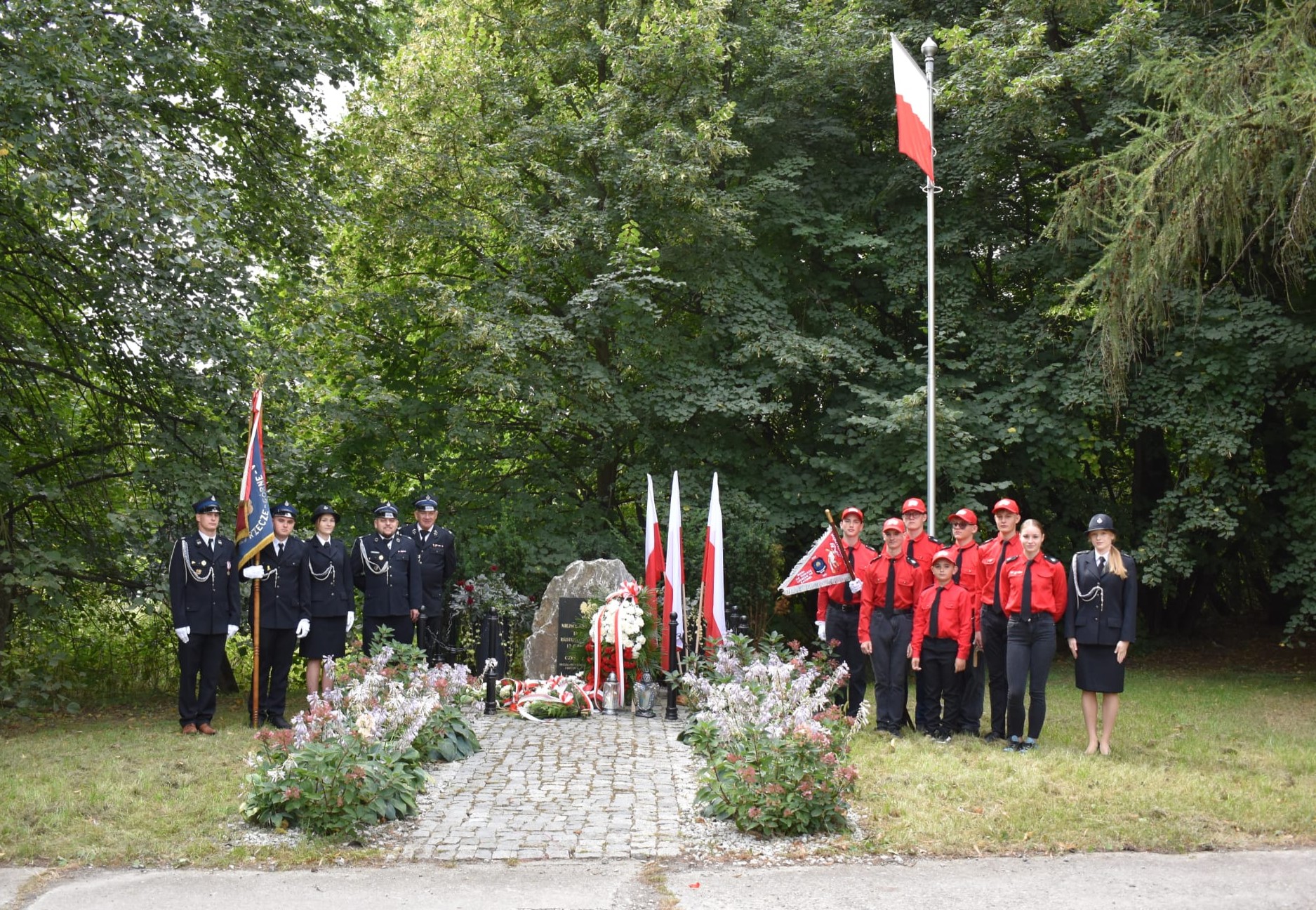 Na zdjęciu widać grupę osób stojących po obu stronach pomnika upamiętniającego, udekorowanego biało-czerwonymi kwiatami i wieńcami. Po lewej stronie stoją osoby w czarnych mundurach galowych strażackich z biało-czerwono-niebieską flagą. Po prawej stronie stoi grupa młodzieży w czerwonych koszulach i czapkach, trzymająca sztandar, obok nich znajdują się dwie osoby w czarnych mundurach. W tle widoczne są trzy polskie flagi ustawione przy pomniku oraz wysoki maszt z powiewającą biało-czerwoną flagą. Scena rozgrywa się w zielonym otoczeniu drzew i krzewów.