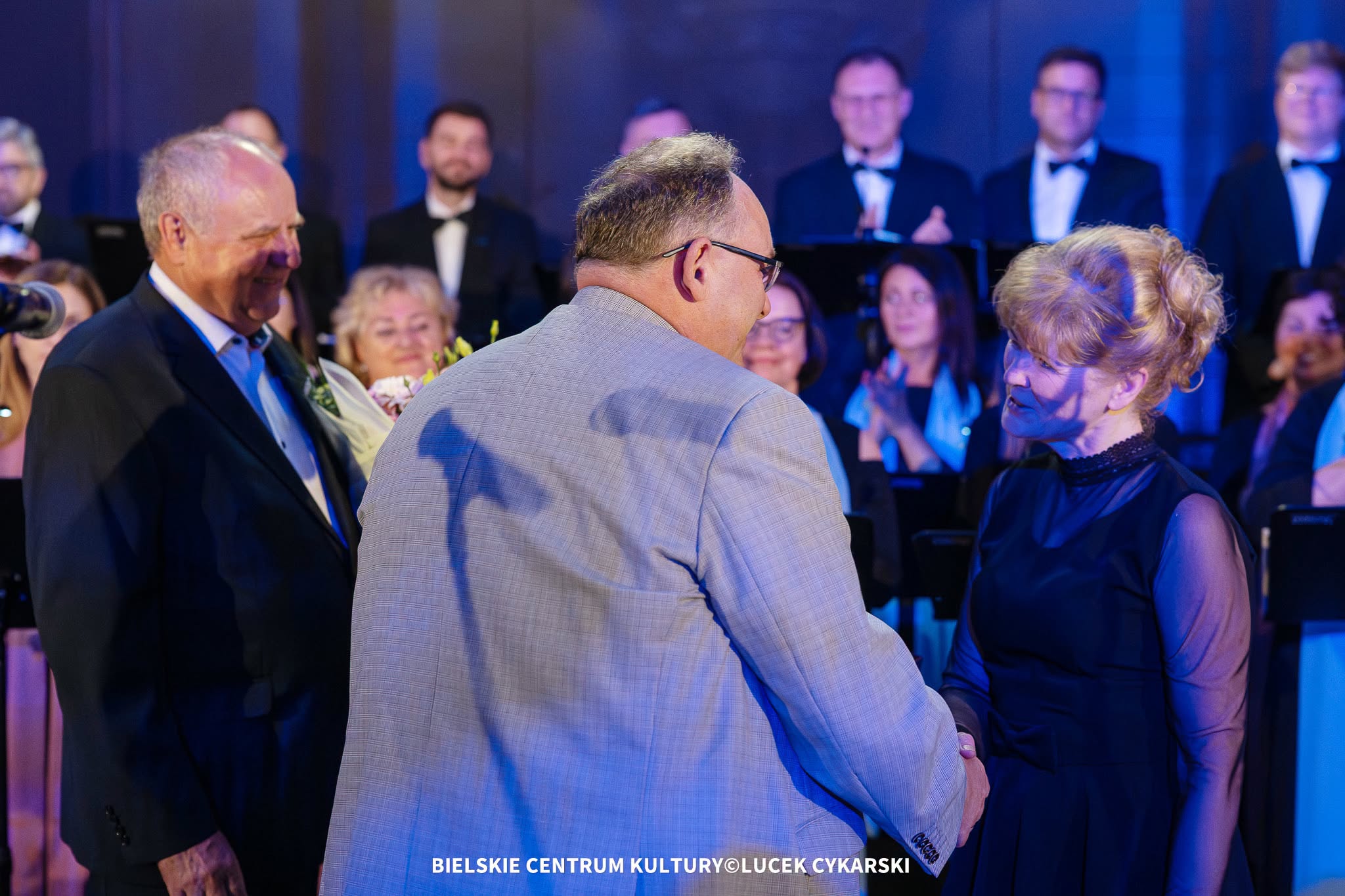 Mężczyzna w jasnej marynarce składa gratulacje kobiecie w granatowej sukni podczas uroczystej gali. Obok stoi starszy mężczyzna z uśmiechem, a w tle widoczny jest chór i publiczność.