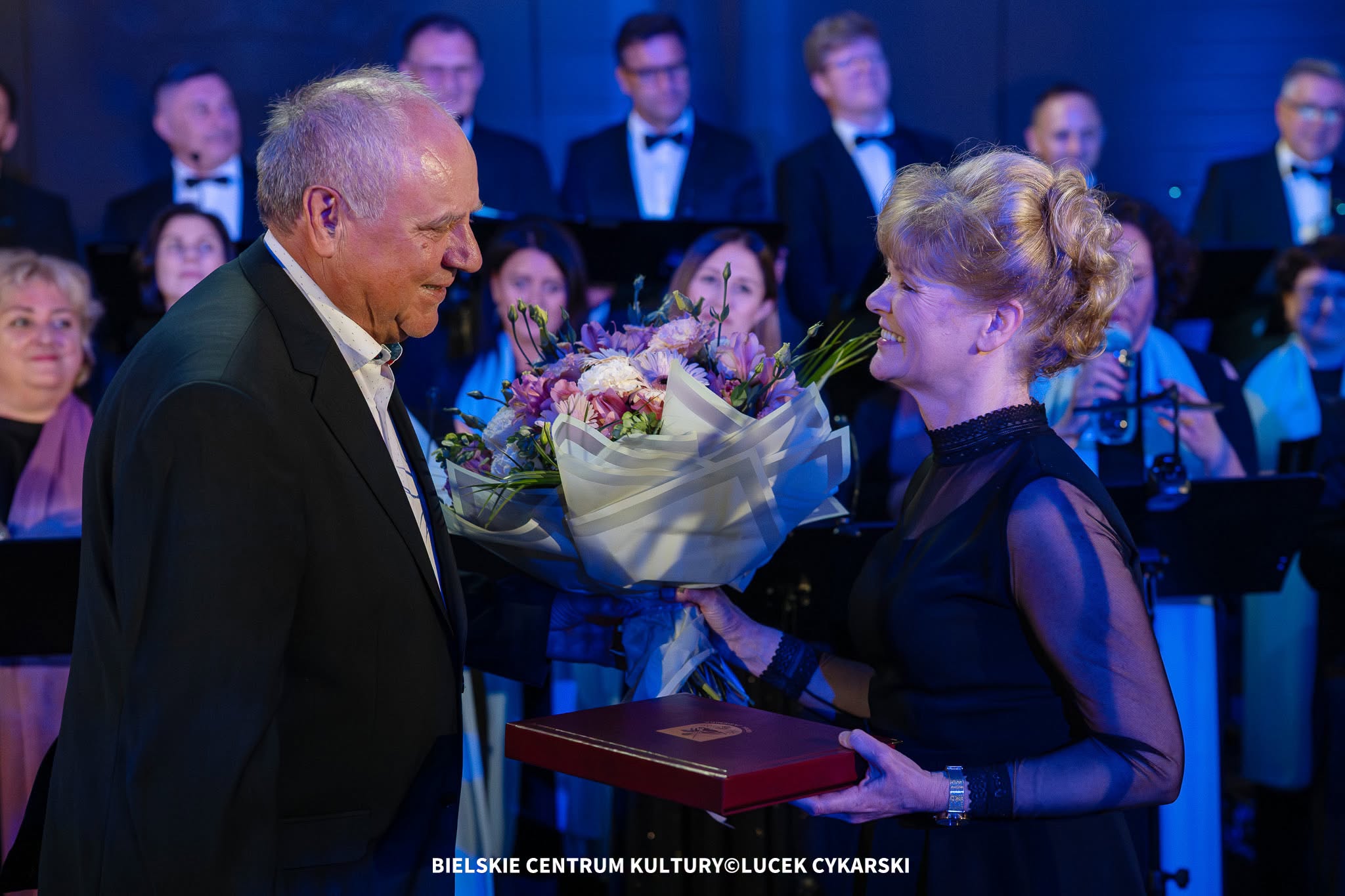 Mężczyzna w jasnej marynarce podczas uroczystej gali wręcza kobiecie w granatowej sukni bukiet kwiatów i pamiątkowe etui podczas uroczystej gali. W tle znajdują się członkowie chóru oraz zgromadzeni goście.