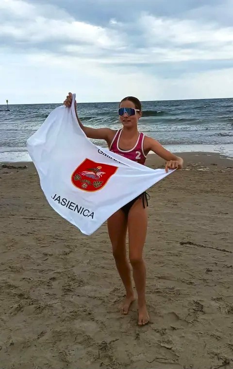 Młoda zawodniczka siatkówki plażowej stoi na plaży nad morzem i trzyma rozłożoną flagę gminy Jasienica.