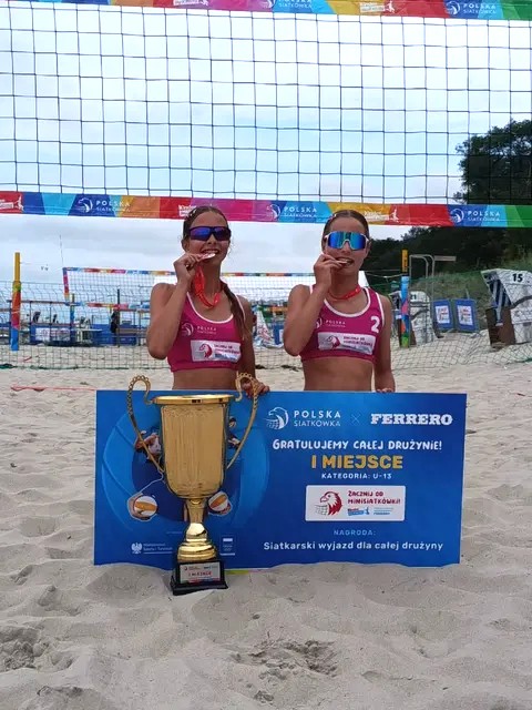 Dwie młode zawodniczki siatkówki plażowej stoją na piasku przed siatką z dużym pucharem i tablicą informującą o zajęciu pierwszego miejsca w kategorii U13.