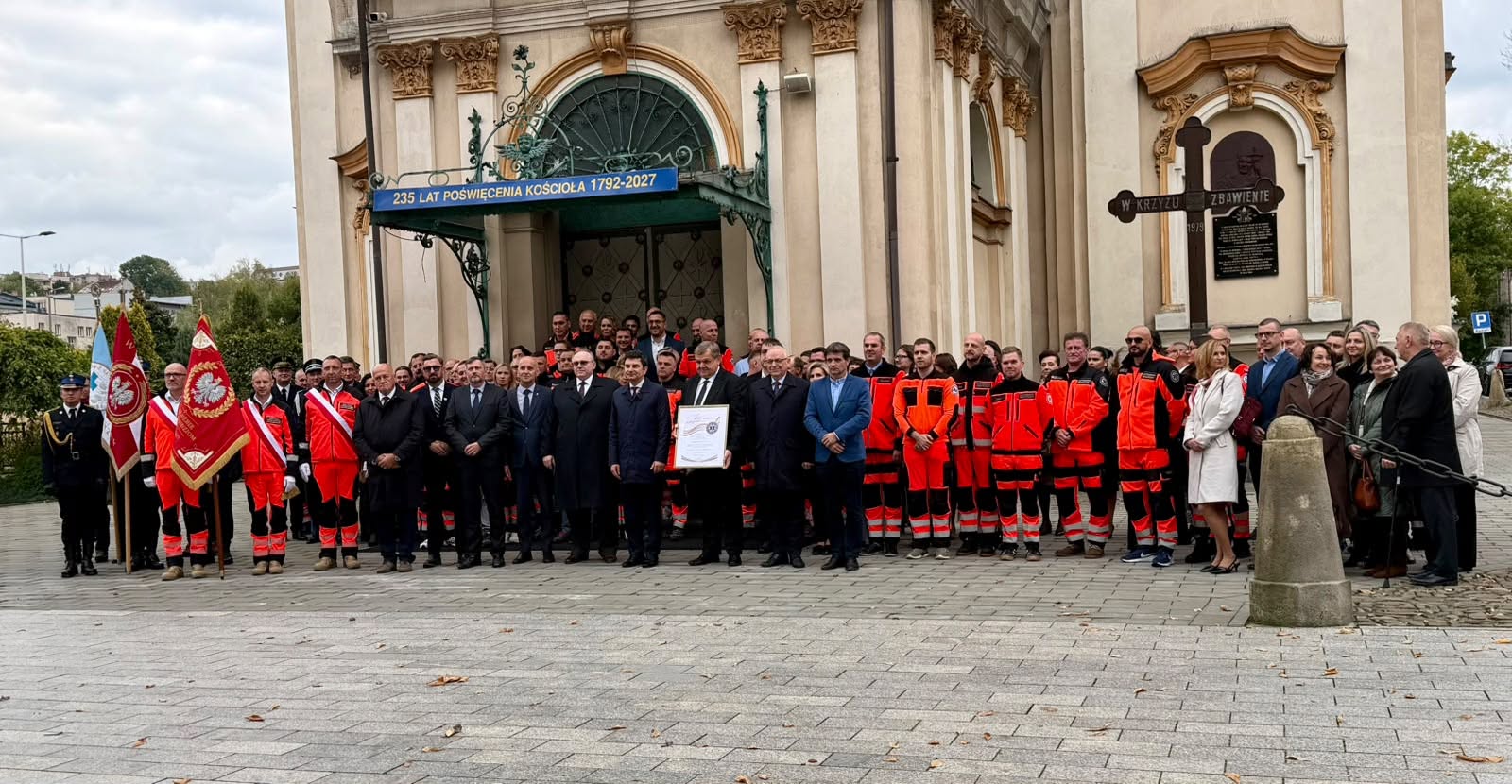 Osoby w oficjalnych strojach, w tym mężczyźni w garniturach, z których jeden dzierży pamiątkowy certyfikat. Po bokach stoją również reprezentanci służb ratowniczych oraz poczet sztandarowy trzymający narodowe flagi.