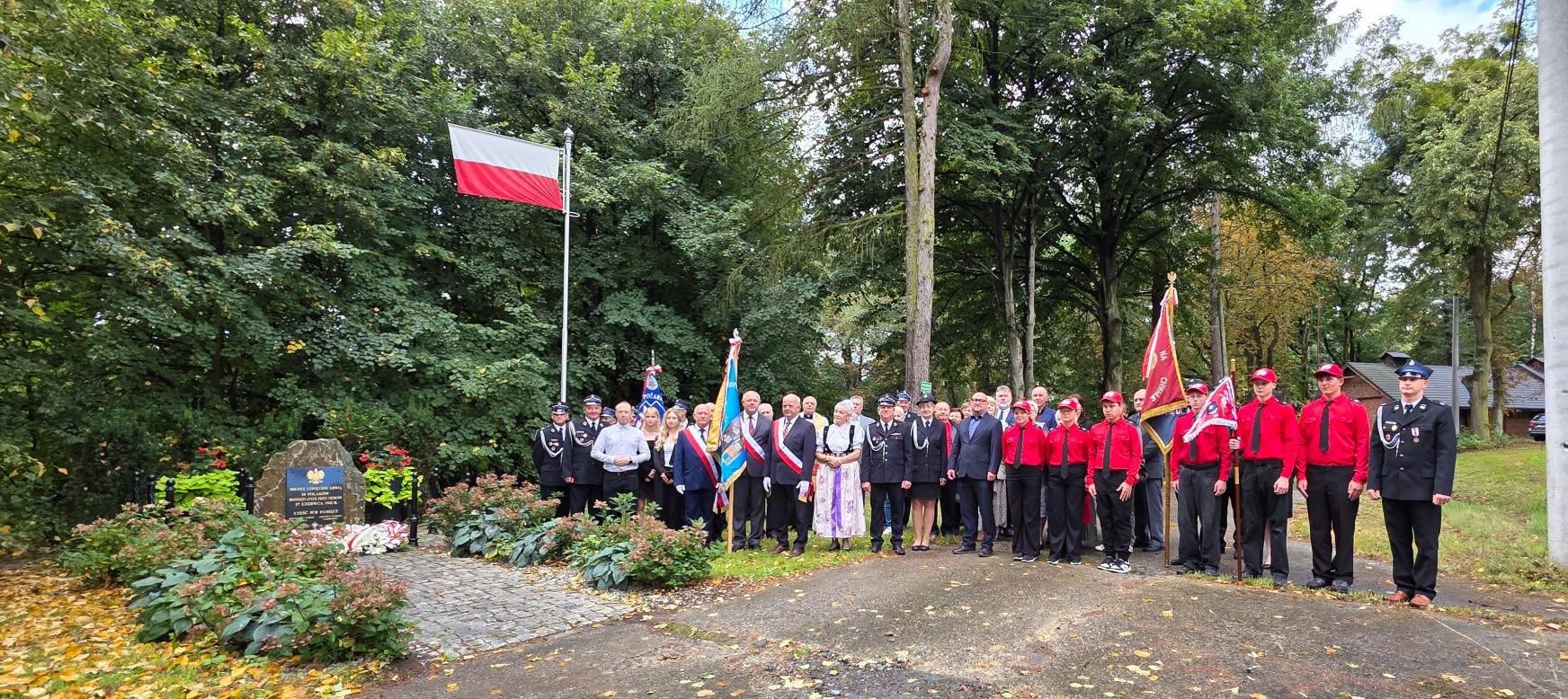 Grupa uczestników uroczystości zgromadzona przy pomniku, wśród nich przedstawiciele straży pożarnej, młodzież oraz osoby z pocztami sztandarowymi. Nad grupą powiewa flaga Polski, a całość znajduje się w otoczeniu parku.