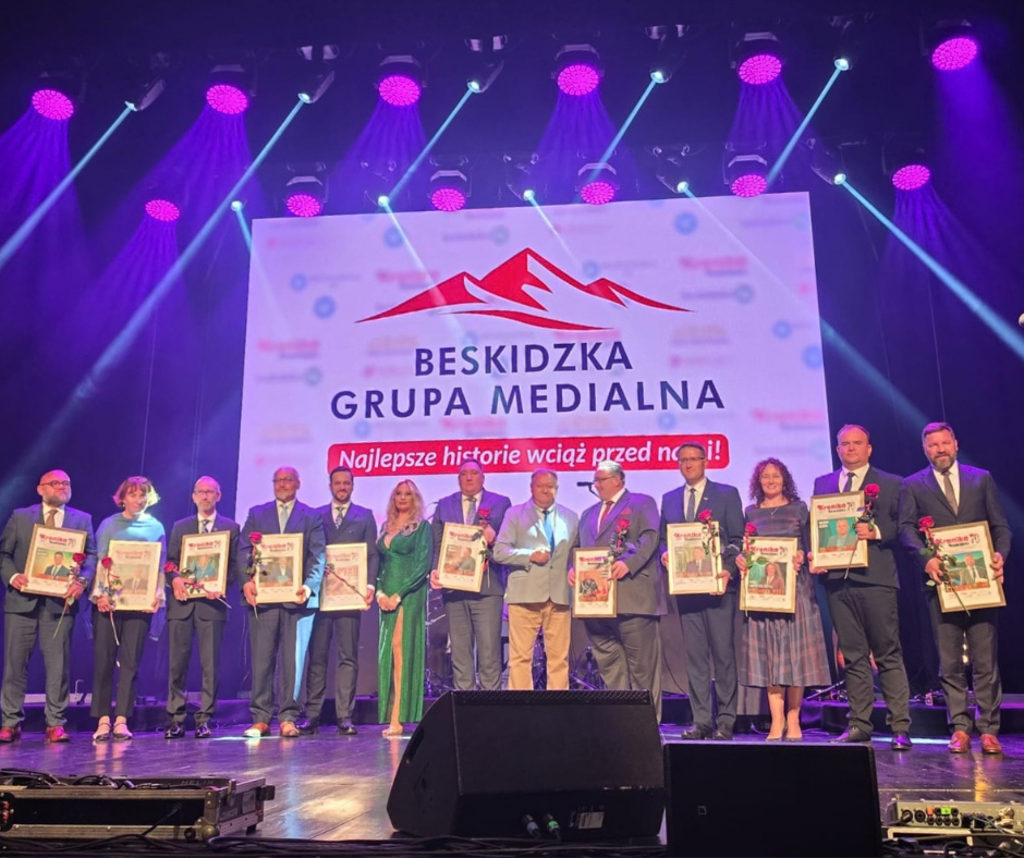 Grupa osób stoi na scenie podczas uroczystości 70 lecia „Kroniki Beskidzkiej”. Uczestnicy trzymają oprawione okładki pisma i kwiaty. W tle widoczne jest logo Beskidzkiej Grupy Medialnej oraz napis „Najlepsze historie wciąż przed nami!”. Nad sceną świecą reflektory w kolorze fioletowym i niebieskim.